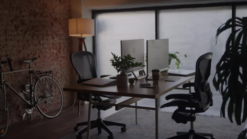 A wellfurnished modern home office space featuring a large wooden desk with two computer workstations and ergonomic chairs situated against an exposed brick wall a large indoor plant and a wide window that provides soft diffused natural light