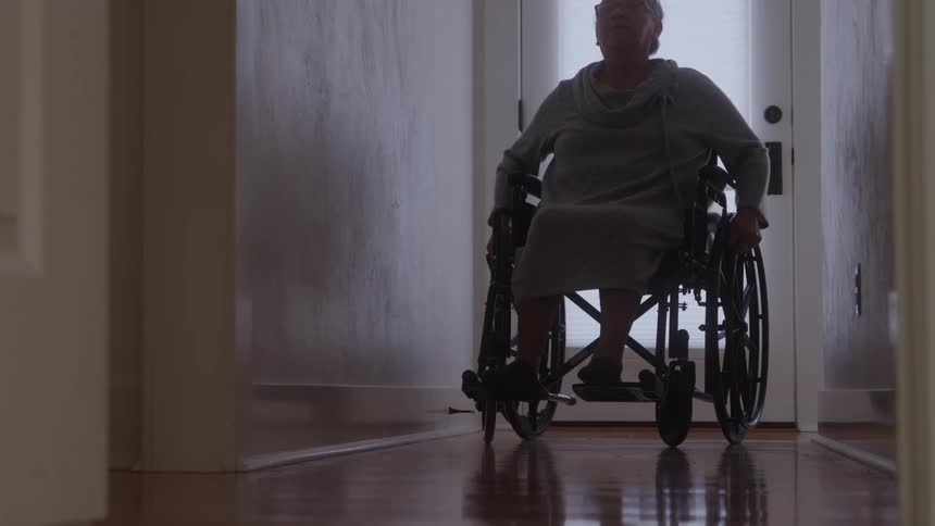 An elderly woman in a wheelchair propels herself down a hallway with a shiny wooden floor moving away from a brightly lit closed door as the shot is framed through a dark foreground doorway
