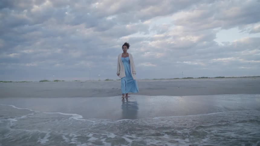 Woman walks toward the water on the beach as her long dress moves in the breeze