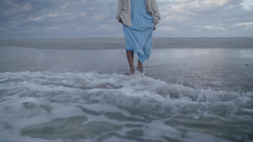 A woman wearing a long light blue dress and a lightcolored cardigan walks barefoot through the shallow water at the edge of the shore with the ocean waves washing over her feet under a cloudy sky