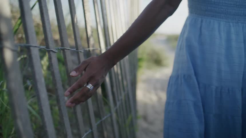Woman walking on a dirt road runs her hand over a wooden fence on a summer day