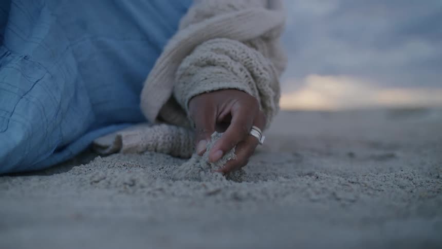 A hand wearing a silver ring gently sifts through the sand on a beach