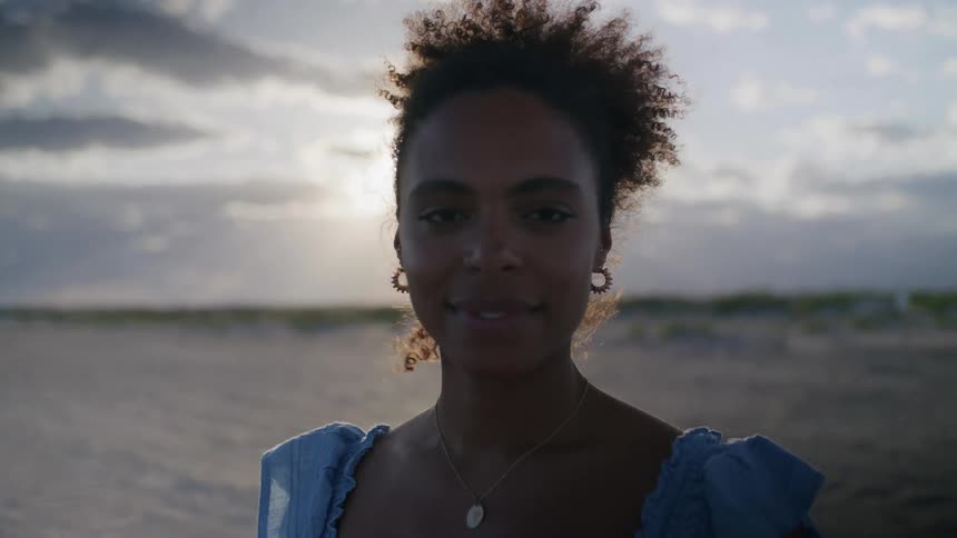 A woman with dark curly hair and a warm smile looks directly into the camera in a closeup portrait backlit by the sun breaking through a cloudy sky on a sandy beach