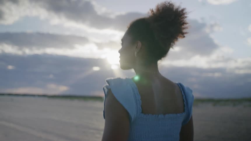 Young black woman wearing a blue dress walking and spining on a beach outdoors during dusk or dawn