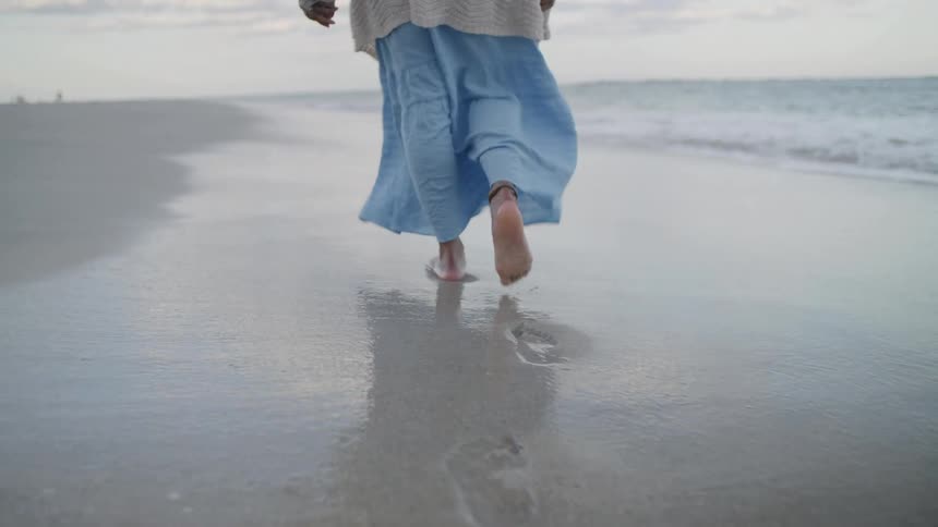 A lowangle shot follows the bare feet and flowing blue pants of a person walking away from the camera along the wet edge of a sandy beach