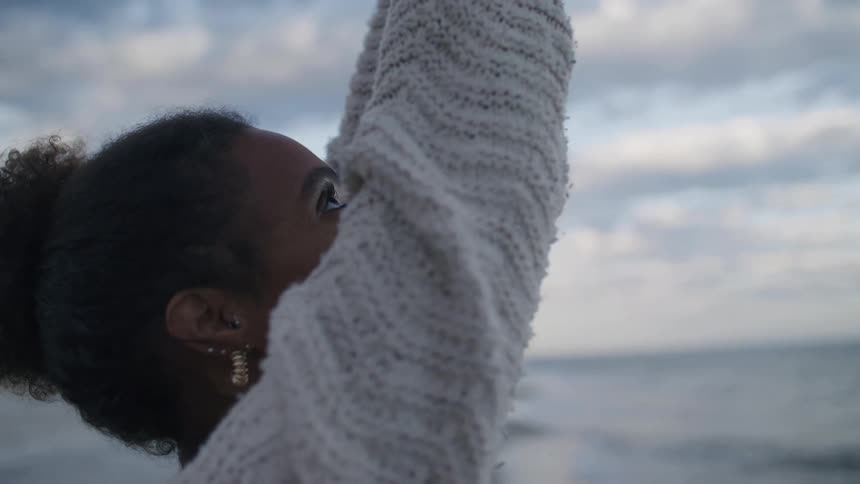 A young woman with curly dark hair wearing a chunky knit sweater and earrings shows her standing on a beach looking up at the cloudy sky as she slowly raises both arms above her head