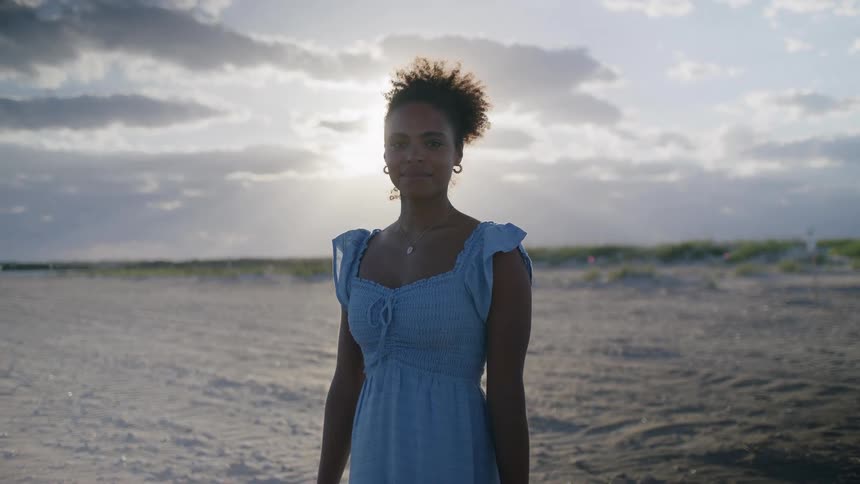 Young black woman wearing a blue dress standing and smiling at the beach during day time