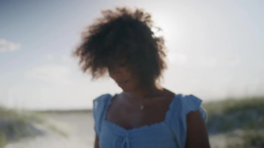 Woman wearing a blue dress examines an object while standing on the beach on a sunny day
