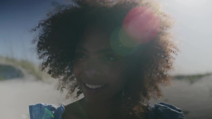 A closeup shot features a young woman with a large afro of dark curly hair smiling genuinely into the camera on a sunny day she is standing outdoors in a sandy possibly beach or dune environment wearing a blue top with ruffled shoulders