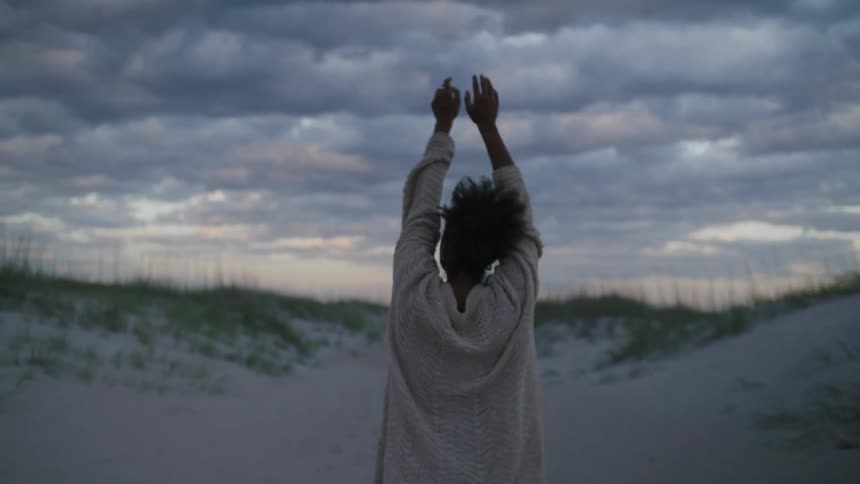 A woman standing on white sand with her back to the camera wearing an oversized white knit sweater raises and stretches her arms wide toward a dramatic cloudy sky