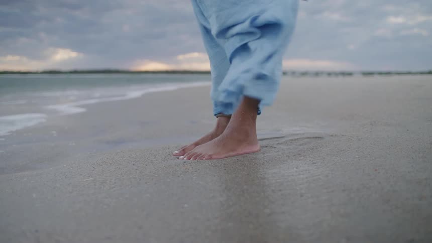 Womans long dress moves in the wind as she stands barefoot on the beach and waves roll in to shore and over her feet