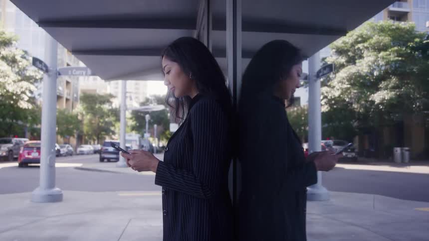 A medium shot of a woman in a dark pinstripe suit standing against a reflective glass panel outdoors while texting on her phone on a city street with traffic in the background