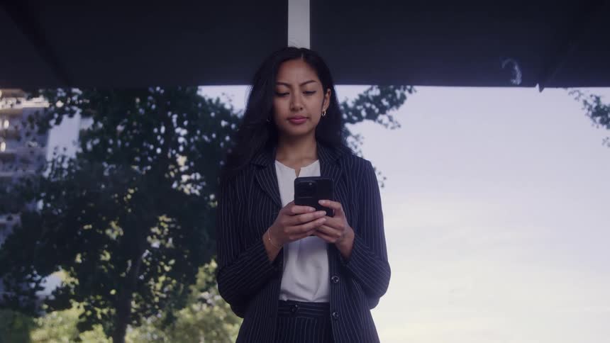 A medium shot of a professional woman in a dark pinstripe suit standing under an awning and typing on her smartphone with green trees visible in the background