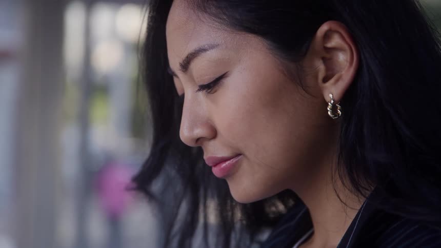 A close profile view of a woman with long dark hair and gold hoop earrings looking down in contemplation in an urban outdoor setting