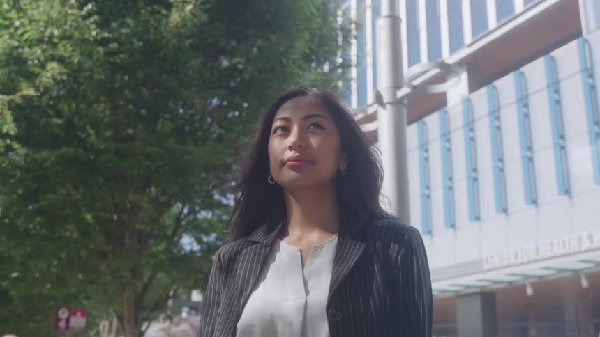 An attractive young woman with long dark hair wearing a white shirt and a pinstripe blazer stands outside on a sunny dayshe looks up thoughtfully with a modern glass building and bright green foliage in the background
