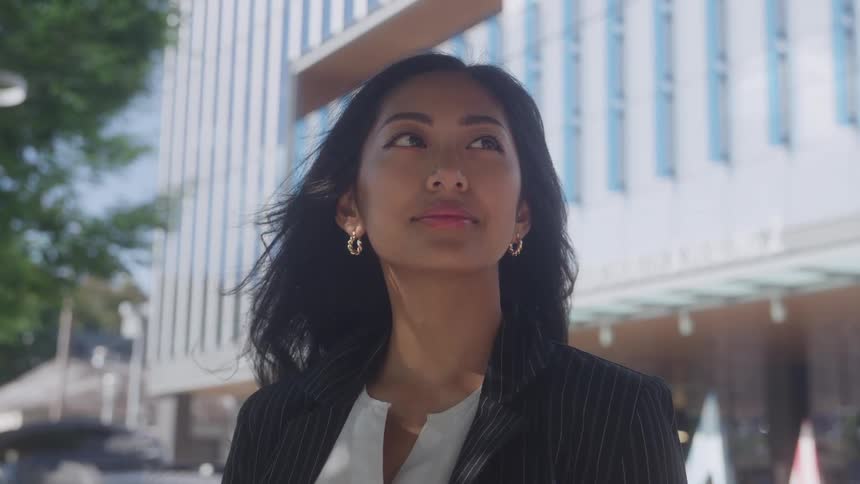 A professional woman with long dark hair wearing a pinstripe blazer and a white shirt captures her walking or standing outdoors in a bright urban area looking upward and offcamera with a determined expression against a modern building