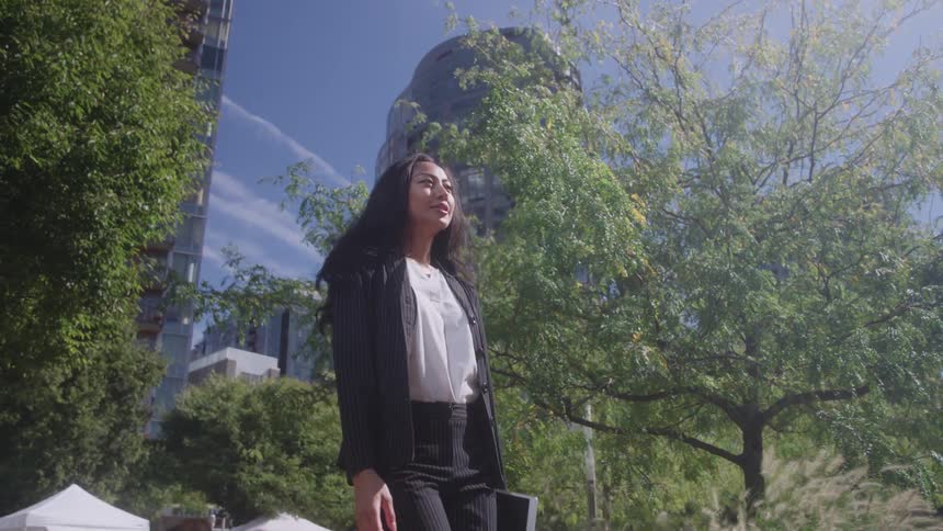 A professional woman wearing a dark pinstripe suit and white blouse is walking confidently through a sunny city park area holding a tablet and looking forward with large buildings and a clear blue sky visible