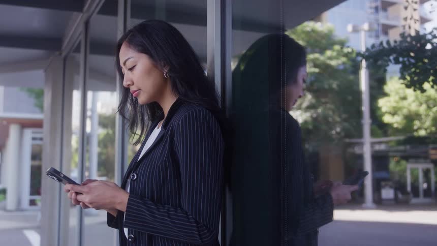 Woman standing outside next to a building texting on her phone with her reflection on the window