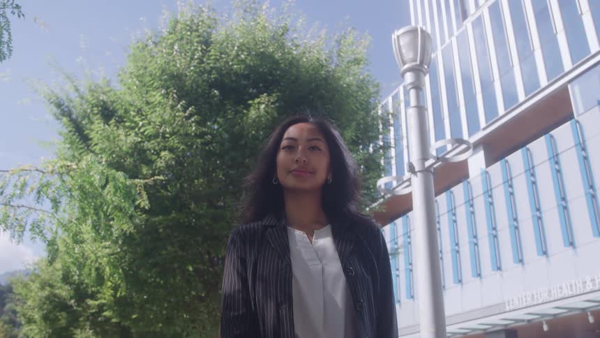 A woman with dark hair stands outdoors with a neutral expression looking forward while wearing a pinstriped jacket against a background of green trees and a modern glass building