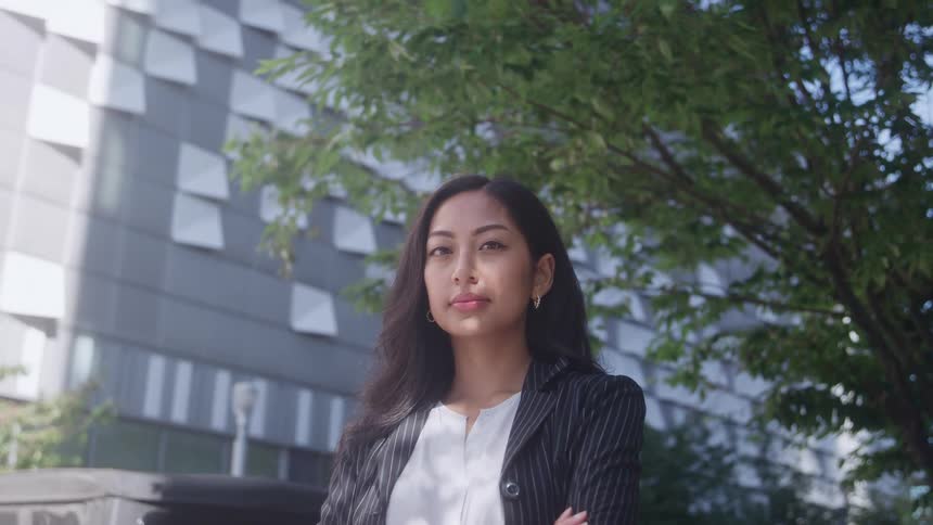 A medium shot of a professional woman in a dark pinstripe suit and white shirt standing outdoors with her arms crossed and eyes closed in front of a modern building and green trees