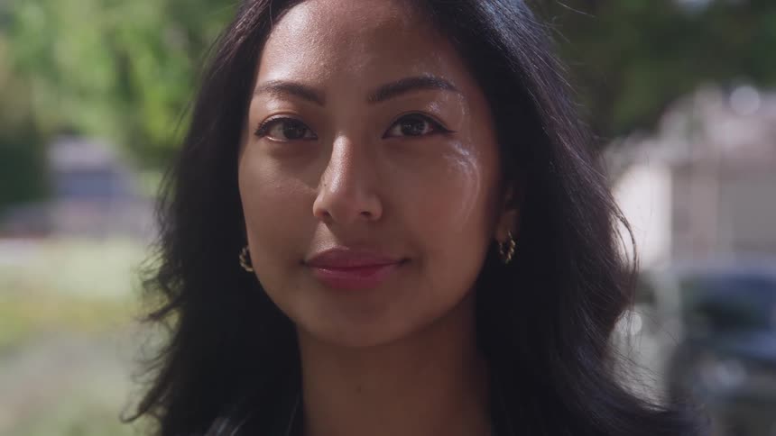 A close view of a beautiful woman with long dark hair opening her eyes and then smiling directly at the camera outdoors on a bright day with greenery in the blurred background