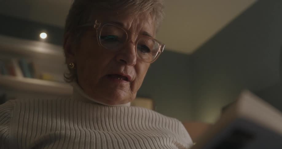 An elderly woman wearing glasses and a white turtleneck is captured in a dark lowangle closeup as she reads a book with the background consisting of a dimly lit interior showing a bookshelf above her