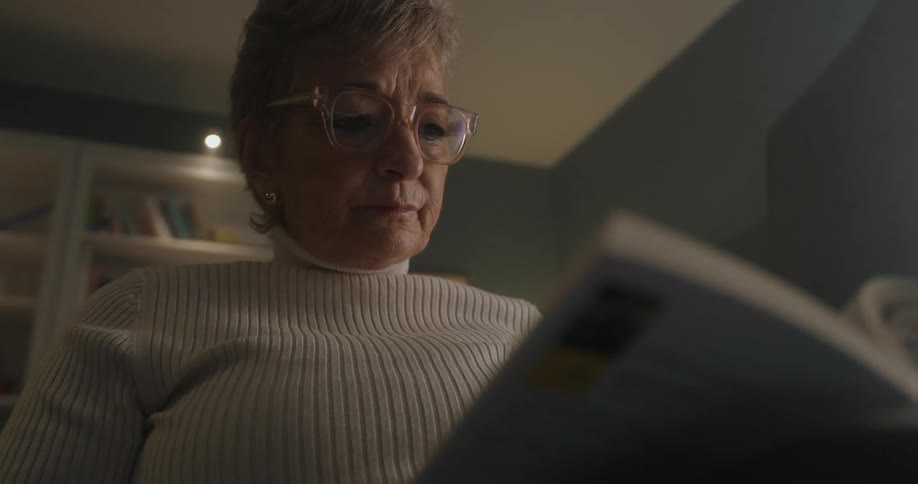 A low angle close view of an older woman with short gray hair and glasses reading a book in a white turtleneck sweater in a dimly lit room with a blurred bookshelf