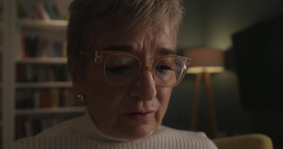 An elderly woman with glasses talks thoughtfully during a video call her expression focused in a warmly lit room