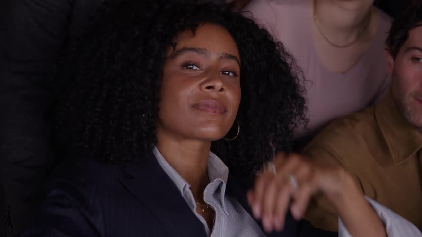 A woman with curly hair smiles gently while seated in a dimly lit room surrounded by others