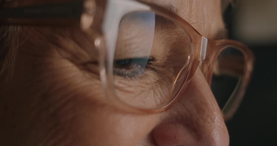 A closeup of an older persons face with a concentrated expression looking forward through large lightcolored framed glasses indicating focus