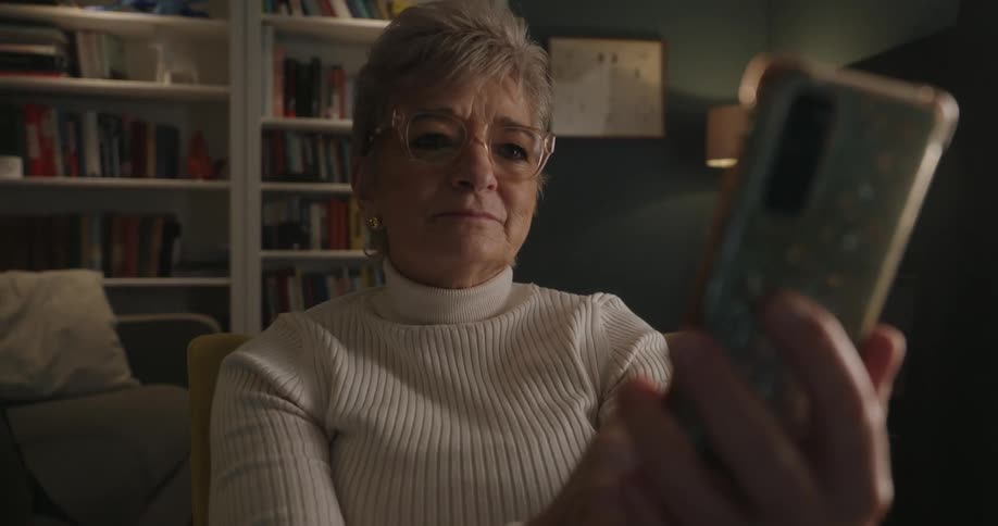 An older woman sits in a chair scrolling on her smartphone in a dimly lit living room