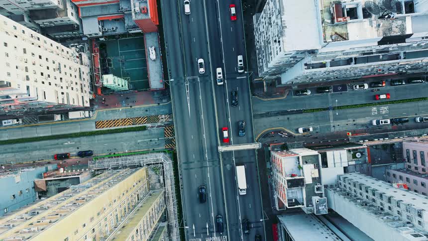 Numerous cars pass along a roadway that is lined by tall buildings