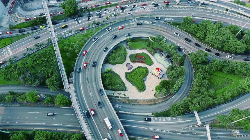 Streams of traffic and bright red taxis flow continuously around a complex circular highway interchange as the camera descends vertically toward the central green park