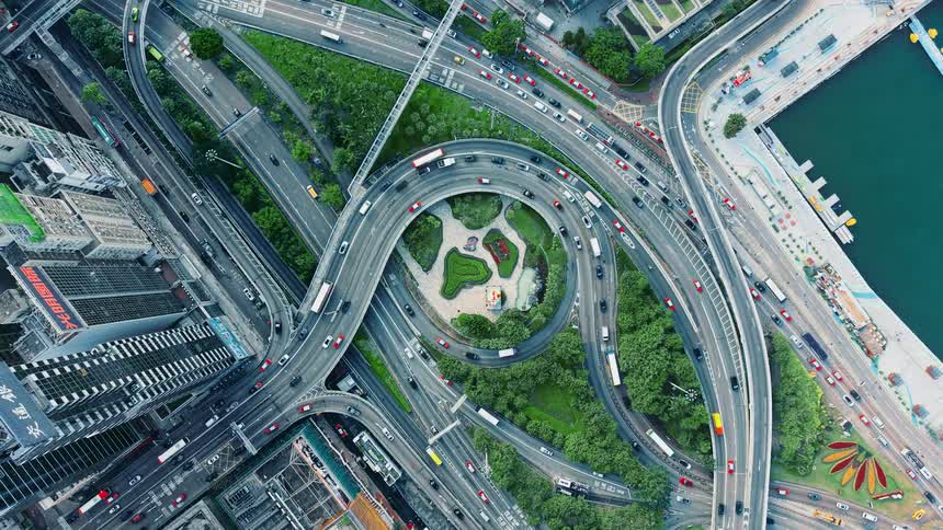 An aerial overhead view captures a complex multi level highway interchange with traffic surrounding a small central green park area near a waterfront