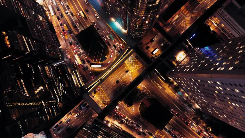 A complex brightly lit urban highway interchange and street intersection at night showing heavy streams of moving and stationary automobile traffic passing between towering skyscrapers