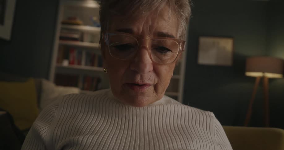 An older woman in a white turtleneck and glasses reads intently looking down at a book in a cozy dimly lit living room