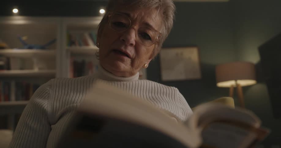 A older woman is sitting in a cozy living room reading out loud from a book