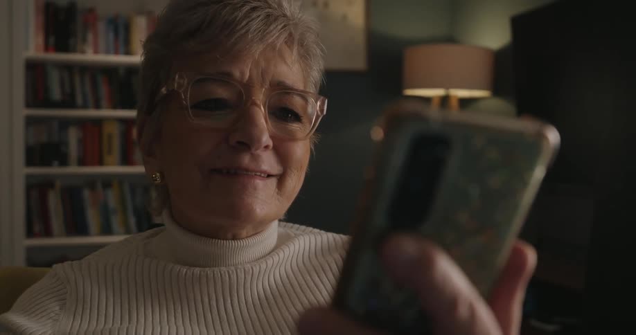 An older woman with short blonde hair and glasses smiles while holding a smartphone in a dimly lit room with a bookshelf and lamp in the background