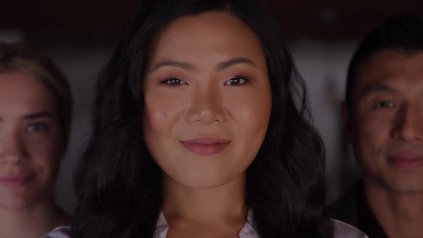 A closeup shot focuses on a woman in the center who turns from a slight profile to face the camera and give a warm smile flanked by the smiling blurred faces of a caucasian woman on her left and an asian man on her right
