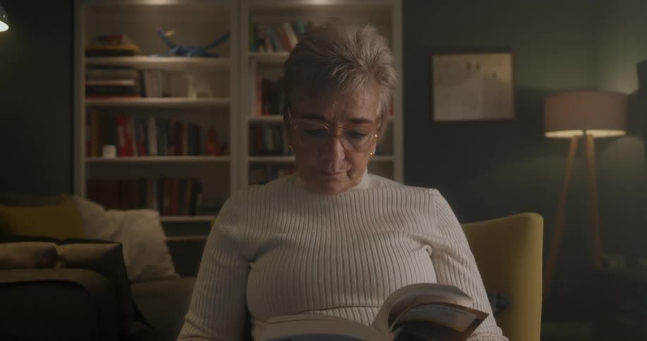 An older woman with blonde hair and glasses wearing a white turtleneck sweater sits reading a book in a comfortable living room with a bookcase behind her