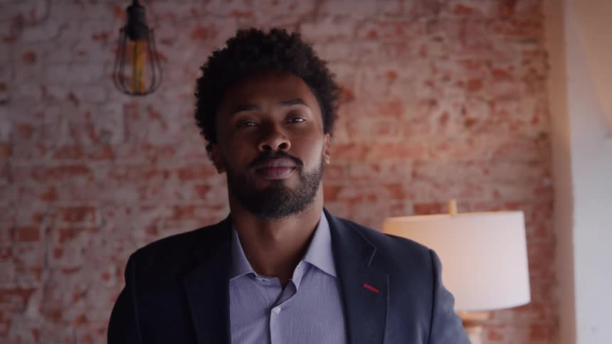 A medium closeup shot focuses on a young black man in a suit jacket and collared shirtwith a confident expression against the backdrop of an exposed brick wall