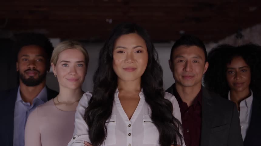 A medium shot of a diverse group of five smiling coworkers with the woman in the foreground looking confidently at the camera with her arms crossed