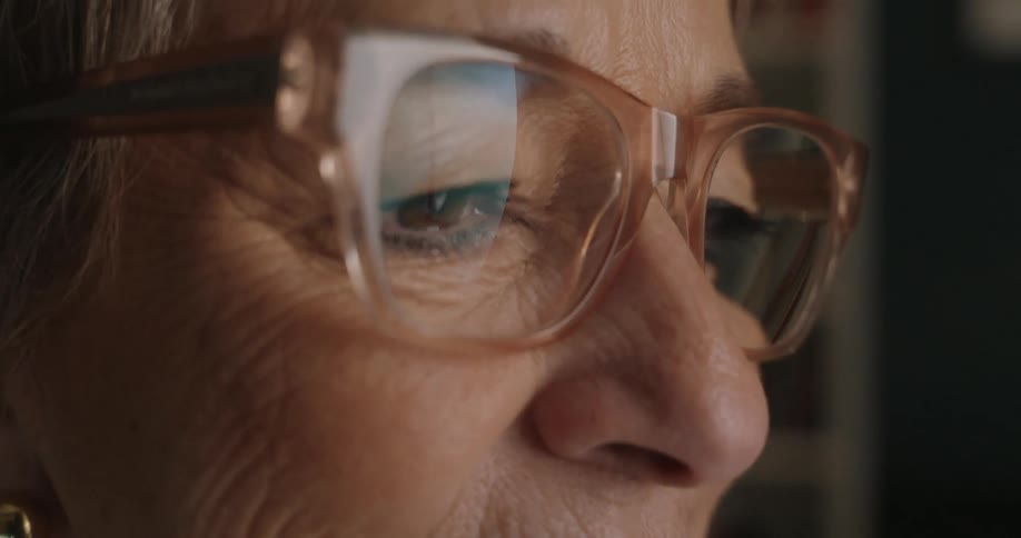An extreme closeup shot focuses on the side profile of an elderly womans eye behind her glasses showing the reflection in the lens as she observes something offcamera with a subtle expression