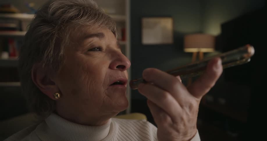 A closeup side profile of an elderly woman shows her holding a smartphone near her mouth and speaking into it while looking upwards in a warmly lit dark room