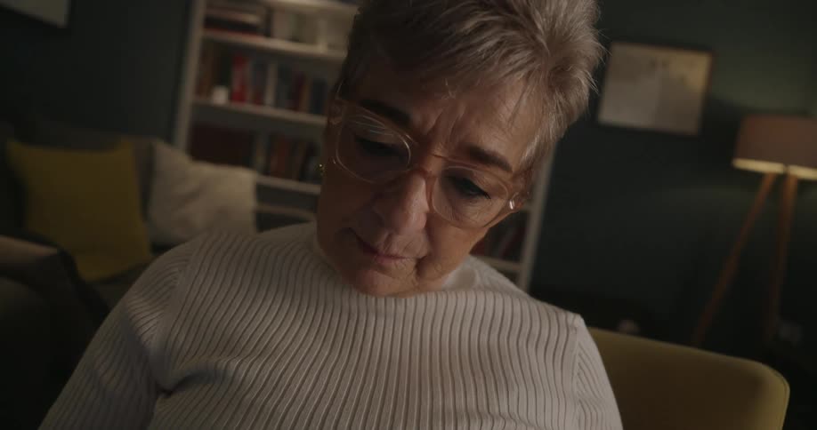 A closeup shot of an elderly woman wearing glasses shows her sitting in a brightly lit armchair against a dark background looking down intently as she reads a physical book