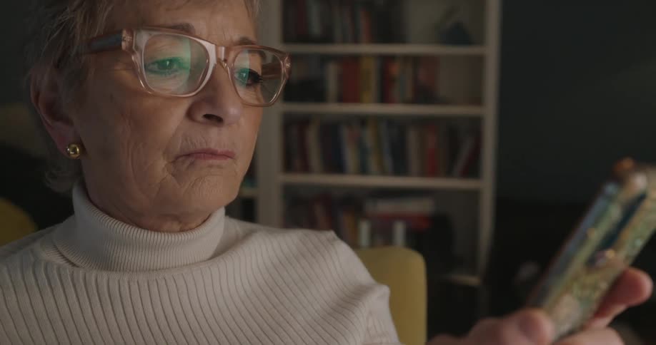An older woman with short gray hair types on her smartphone while seated in a dimly lit book filled room