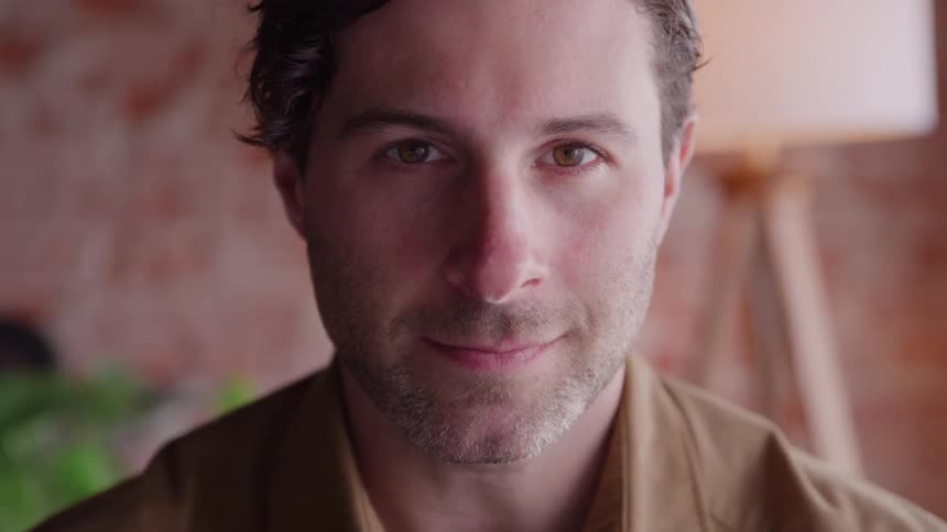 A man with brown hair and light stubble looks directly into the camera with a calm friendly expression and a slight subtle smile in a warmly lit indoor setting