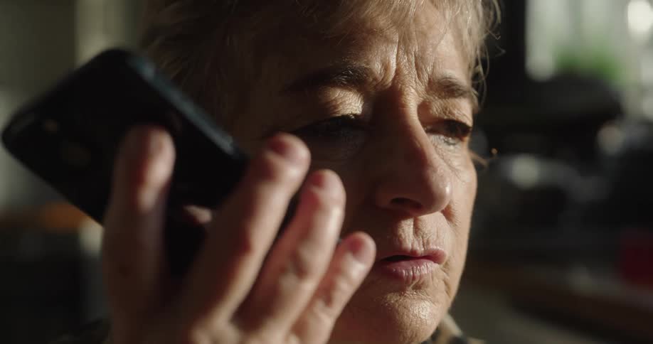 A close view of an older woman with short hair holding a smartphone to her mouth while talking seriously in soft indoor lighting