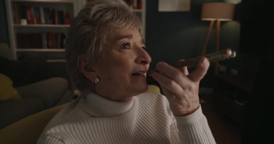 An older woman with short gray hair is sitting in a comfortable chair and using a smartphone on speaker to talk or send a voice message she is wearing a creamcolored turtleneck sweater and smiling brightly as she speaks into the phone