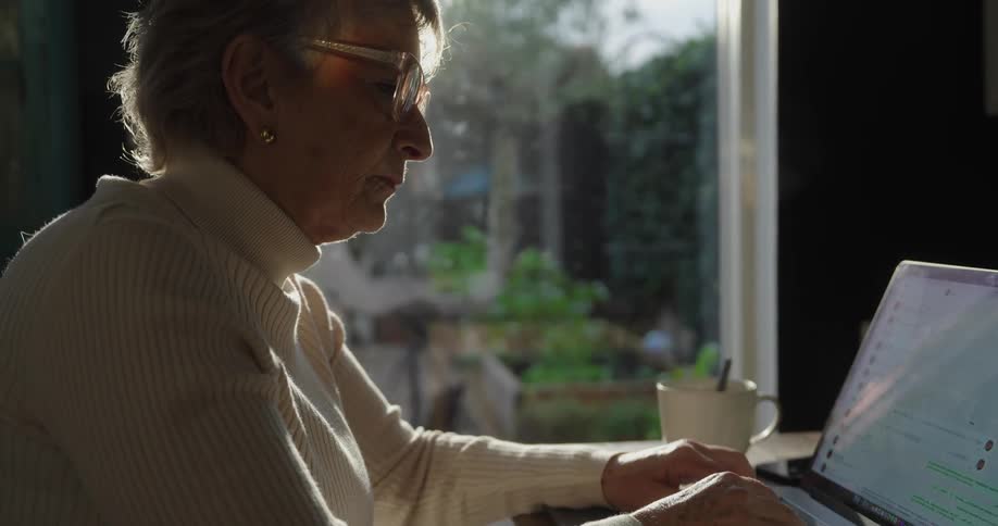 An elderly woman dressed in a white turtleneck and wearing glasses concentrating intently while typing on a laptop with her workspace brightly backlit by natural light streaming in through a window that reveals a blurred view of green outdoor foliage